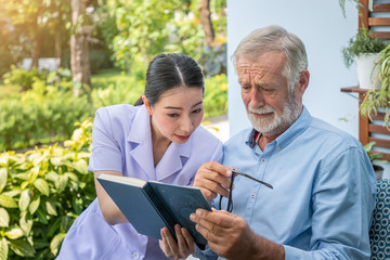 Senior elderly man reading book with nurse during breakfast in garden at nursing home