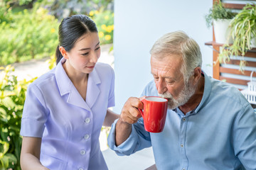 Nurse assist elderly senior man to drink coffee with mug in hand at nursing home