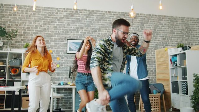 Bearded guy is dancing in office while coworkers are clapping hands having fun together at corporate party. People, lifestyle and modern youth concept.
