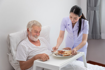 Happy nurse serving elderly senior man breakfast on bed in bedroom at nursing home