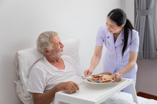 Happy nurse serving elderly senior man breakfast on bed in bedroom at nursing home