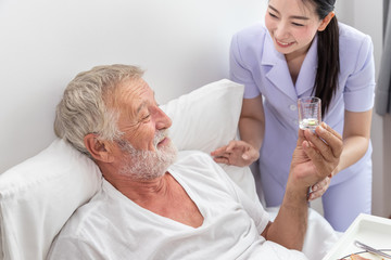 Happy nurse give pills, assist, and encourage elderly senior man to eat medicine after breakfast on bed in bedroom at nursing home