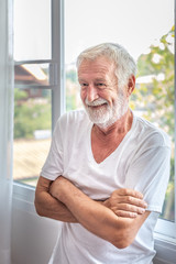 Senior elderly man standing at window in bedroom after waking up in morning, cross arm
