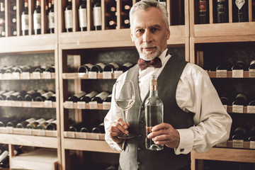 Sommelier Concept. Senior man standing with bottle and glass of white wine smiling joyful