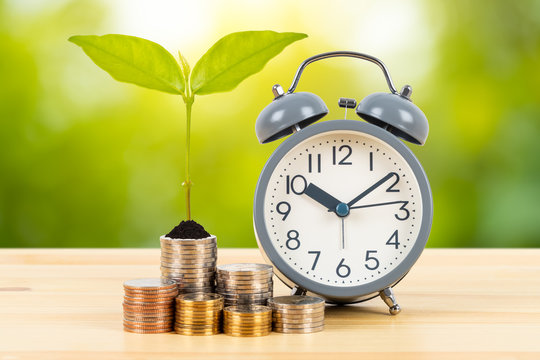 Coin stack with growing leaves and alarm clock on wooden desk  on green tree background, time for saving concept