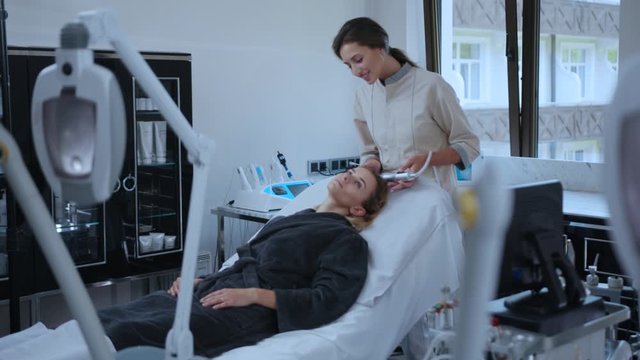 Professional beautician doing rf-lifting cosmetic massage on woman head. Relaxed girl with closed eyes enjoying spa facial treatment in a salon.