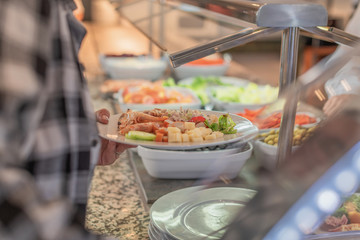 A white plate with shrimp and vegetables held by a woman's hand next to the window of a food buffet. Concept of gastronomic food.