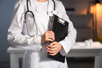 cropped view of doctor in white coat with stethoscope holding clipboard during night shift