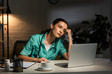 attractive and tired nurse in uniform sitting at table and looking away during night shift