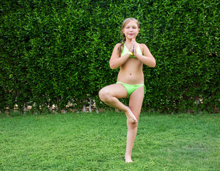 young sports blonde kid girl in green swimsuit is doing yoga exercise on the grass and smiling happy with hands in front of on the green plant wall background. childhood sport concept, free space - obrazy, fototapety, plakaty