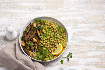 Stewed chickpeas in green sauce with with baked eggplant, parsley and lemon on white wooden background, rustic style, top view. delicious healthy homemade food	