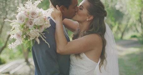 Happy multi ethnic couple kissing on their wedding day with a flower bouquet and green trees in the background with afternoon sun rays filtering through, the perfect wedding day