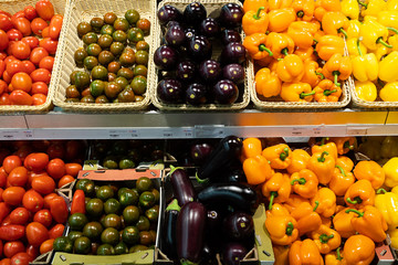 Supermarket counter with wicker baskets and lug boxes with red and green tomatoes, orange and yellow peppers and eggplants