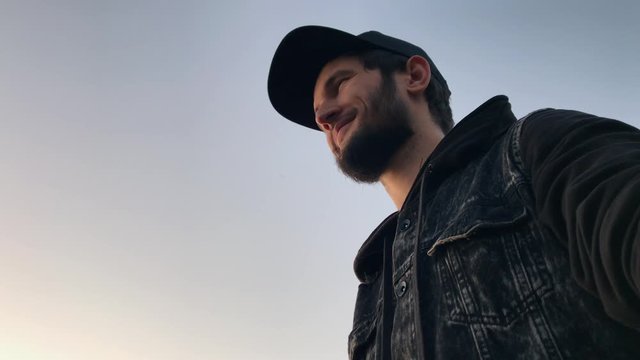 Portrait of young bearded guy wearing black cap and jeans jacket, on background of sky.