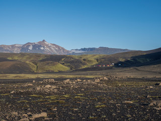 Fototapeta premium View on mountain hut at Iceland on Laugavegur hiking trail, green valley in volcanic landscape among lava fields. Early morning, summer blue sky