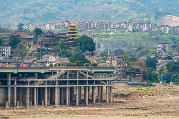 Obraz premium Chinese touristic quarter on the river bank. Against the backdrop residential areas on the hills. Father and son fly a bright red kite on the dry bank of the Jialing River. Chongqing, China.