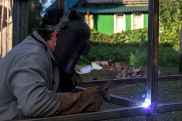 A strong man is a welder, welding mask and welders leathers, a metal product is welded with a arc welding machine at the construction site, blue sparks fly to the sides