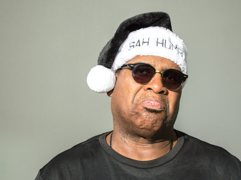 grumpy African American man wearing sunglasses and a black and white Bah Humbug hat isolated on a solid background