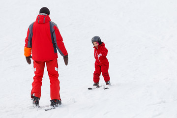Fototapeta na wymiar Little girl in red learning to ski with the help of an adult