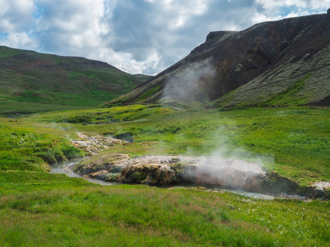 Reykjadalur valley with hot springs river and pool with lush green grass meadow and hills with geothermal steam. South Iceland near Hveragerdi city. Summer sunny morning, blue sky.