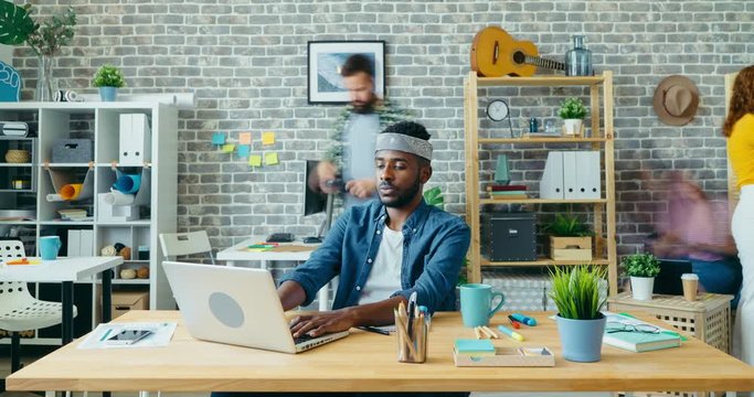 Zoom-in time-lapse of African American man office worker using laptop at work sitting at desk while colleagues men and women moving in background.