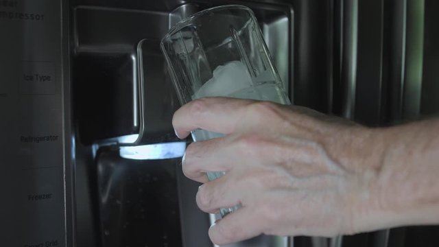 Man filling drinking glass with fresh water from refrigerator purified water dispenser. Stainless steel kitchen appliance with built in water filter.