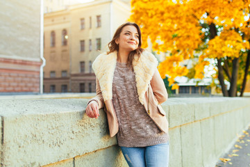 Beautiful caucasian brunette girl standing warm autumn day with background of trees with yellow foliage and a city.