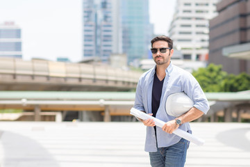 The young handsome man engineer is standing in control and holding a white helmet and construction plan, with a backdrop of a tall building in the morning in Bangkok Thailand. with copy space