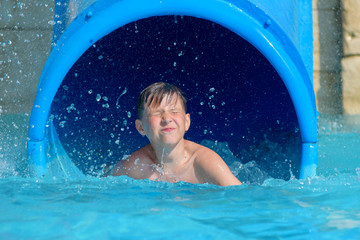 Boy having fun on water slide in aqua fun park glides, happy falling into water and water splashes...