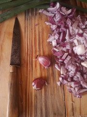 Vista aerea de un cuchillo con dos ajos morados y cebolla roja recién picada sobre una tabla rústica.