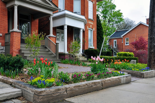 This beautiful urban front yard garden features a large veranda, brick paver walkway, retaining wall with plantings of bulbs, shrubs and perennials for colour, texture and winter interest.