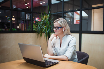 young blonde woman sits at her laptop and uses wireless headphones.