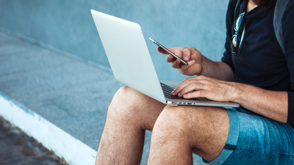 A young man, sitting on a skateboard, is trying to synchronize a laptop with a phone. Look into the frame.