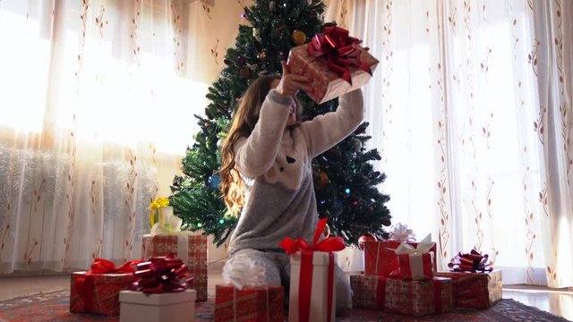 Happy smiling girl with curly hair sitthing on the ground under the christmas tree and looking over the christmas presents. Red boxes with colorful ribbons full of special gifts. Christmas gift boxes