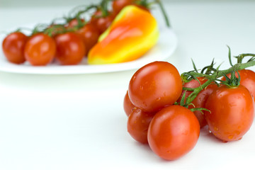 Bunch of red cherry tomatoes on the front and a bunch of fresh red cherry tomatoes and sweet papper on the white plate in the background. Close up view, white background