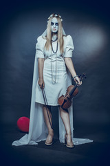 A young girl in a white dress and makeup for Halloween holds a violin in her hands