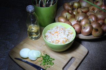bowl of pickled cabbage with green and white onion and oil on wooden cutting board  with knife, ingredients on sackcloth background