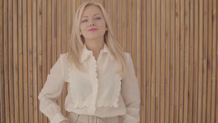 Gorgeous Caucasian woman with blond hair and grey eyes looking at her watch, at the camera and smiling sceptically. Confident young lady posing on the background of wooden wall.