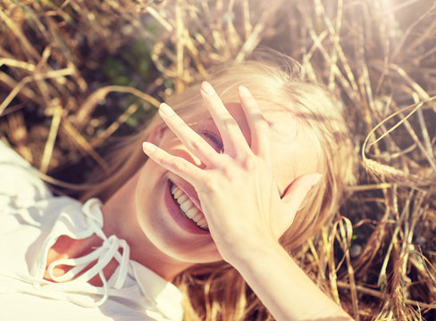 nature, summer holidays, vacation and people concept - close up of happy young woman lying on cereal field and covering face by hand