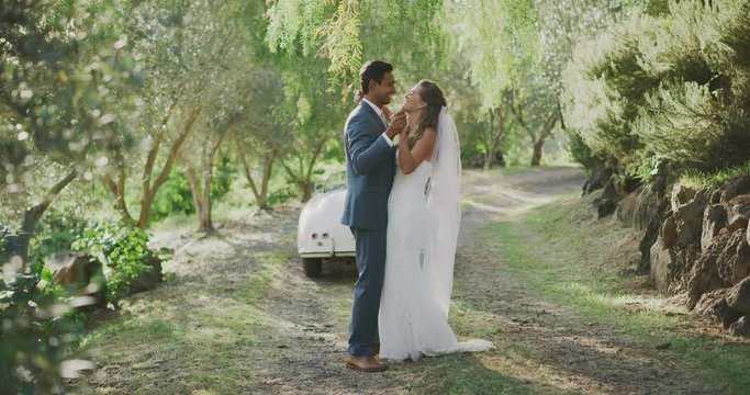 Diverse bride and groom slow dancing in the countryside outdoors on their wedding day, happy multi ethnic married couple slow dancing in the afternoon sunlight with a vintage car in the background