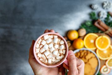 Winter drink. Flat lay of man hands holding a mug with hot chocolate or cocoa and marshmallow, Gingerbread cookie, Cinnamon sticks, cones, sprigs of spruce, mandarin on black background. Copy space