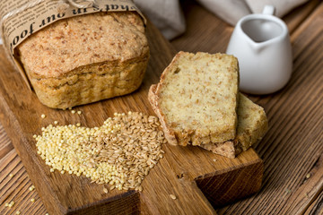 healthy content, a brick of freshly baked golden bread, couple of cut pieces, millet, flax, a scattering of grain, paper wrapper, wooden Board, textile, brown background, horizontal