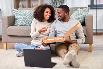 leisure, people and food concept - happy african american couple with laptop computer eating takeaway pizza at home