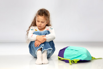 childhood, school and education concept - sad little girl with backpack sitting on floor over grey...