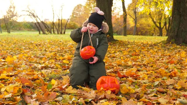 Little girl sway small Jack-O-lantern, sitting at autumn park. Baby play with traditional Halloween decoration. Fallen leaves around, ground covered by yellow maple foliage