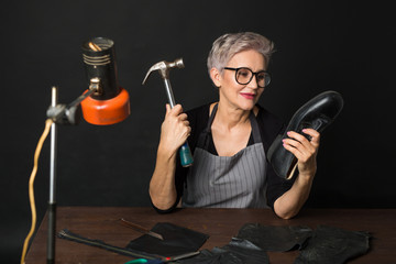 stylish aged woman with glasses in an apron on a black background with a hammer and a boot in her hands