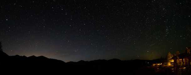 Shooting Star over cabin in the woods lit against starry night sky and shadowed mountain peak backdrop with distant light glowing beyond mountain panoramic landscape