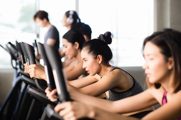 A beautiful Asian Thai woman wearing exercise clothes is riding a bicycle with a backdrop of a group of male and female athletes exercising in the fitness room in Bangkok in the morning.