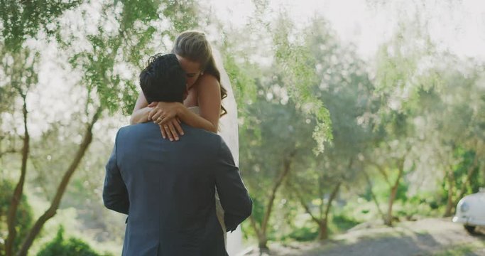 Happy young multi ethnic groom kissing his bride in his arms on their wedding day in nature smiling at each other, happy diverse married couple embracing and kissing in nature