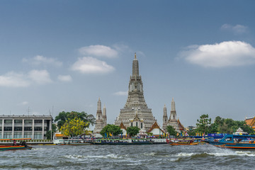 Bangkok city, Thailand - March 17, 2019: The spires or Prangs of Temple of Dawn against blue sky with some white clouds. Some green foliage, Long blue boat on Chao Phraya River up front.